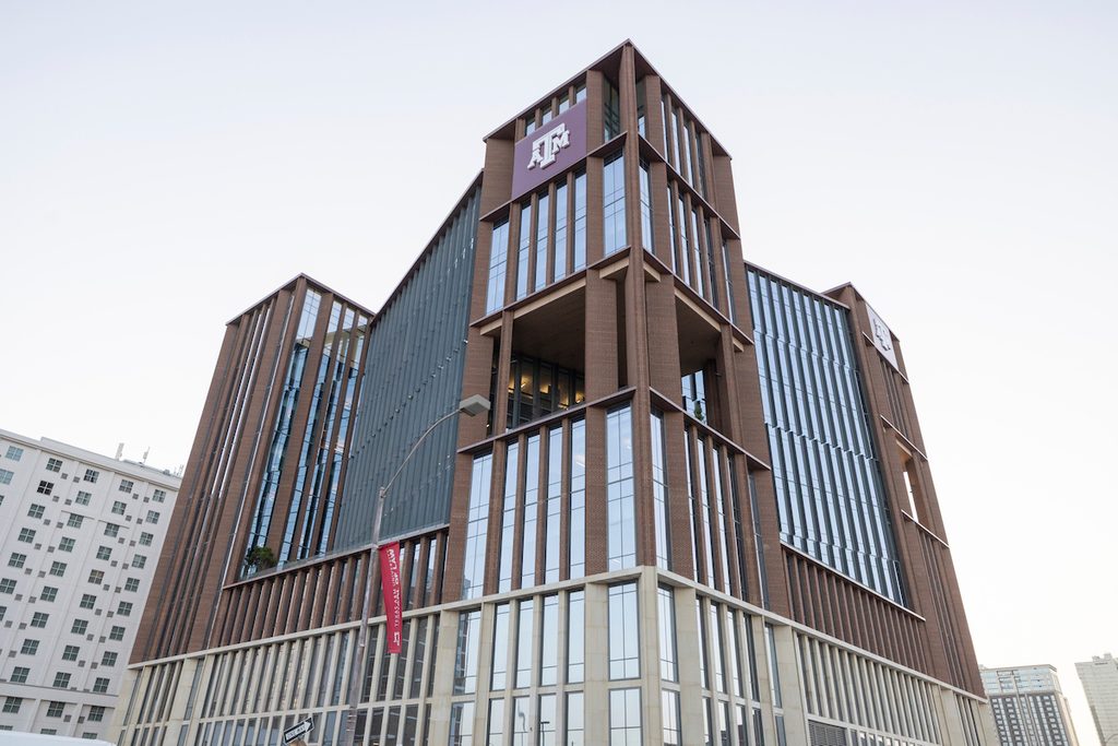 A modern multi-story building with a brown and glass facade features a prominent logo at its peak.